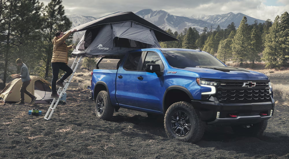 Person setting up a tent over the bed of a blue 2026 Chevy Silverado 1500 parked off-road.