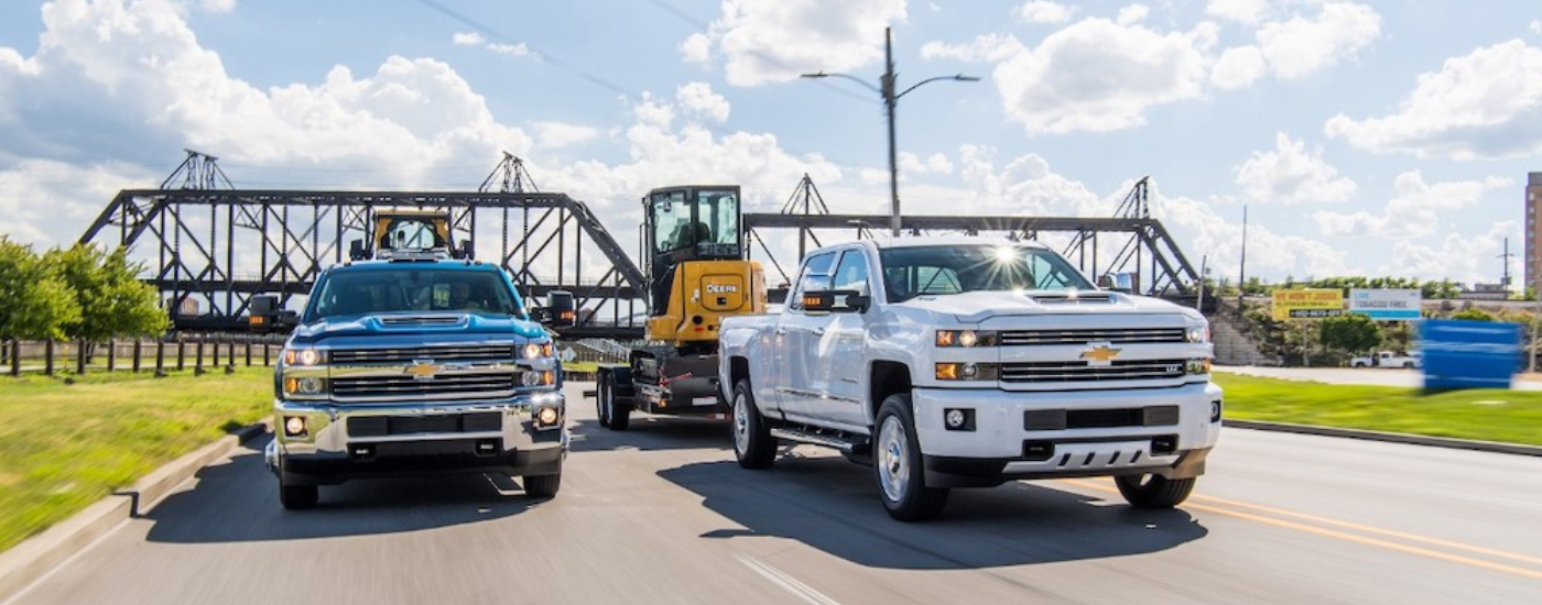 A white and a blue 2018 Chevy Silverado 2500 HD driving side by side towing machinery 